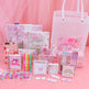 A pink stationery gift set arranged on a pink surface with a pink draped backdrop. The set includes notebooks, pens, a pencil case, sticky notes, stickers, a small card holder, and a large translucent tote bag, all in pastel pink tones with cute cartoon and rainbow designs.