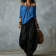 A woman modeling a loose off-the-shoulder blue top paired with voluminous black wide-leg pants, accessorized with layered necklaces, bracelets, hoop earrings, and a woven brown bag, set against a textured gray studio backdrop.