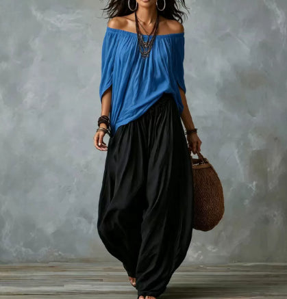 A woman modeling a loose off-the-shoulder blue top paired with voluminous black wide-leg pants, accessorized with layered necklaces, bracelets, hoop earrings, and a woven brown bag, set against a textured gray studio backdrop.