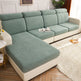 A light sage green textured sofa slipcover fitted on a sectional sofa with cream-colored arms and base, shown in a bright living room setting.