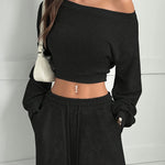 Person wearing a black off-shoulder top and black pants against a white wall.