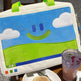 Children's drawing board with a smiley face design on a table with a muffin and drink.