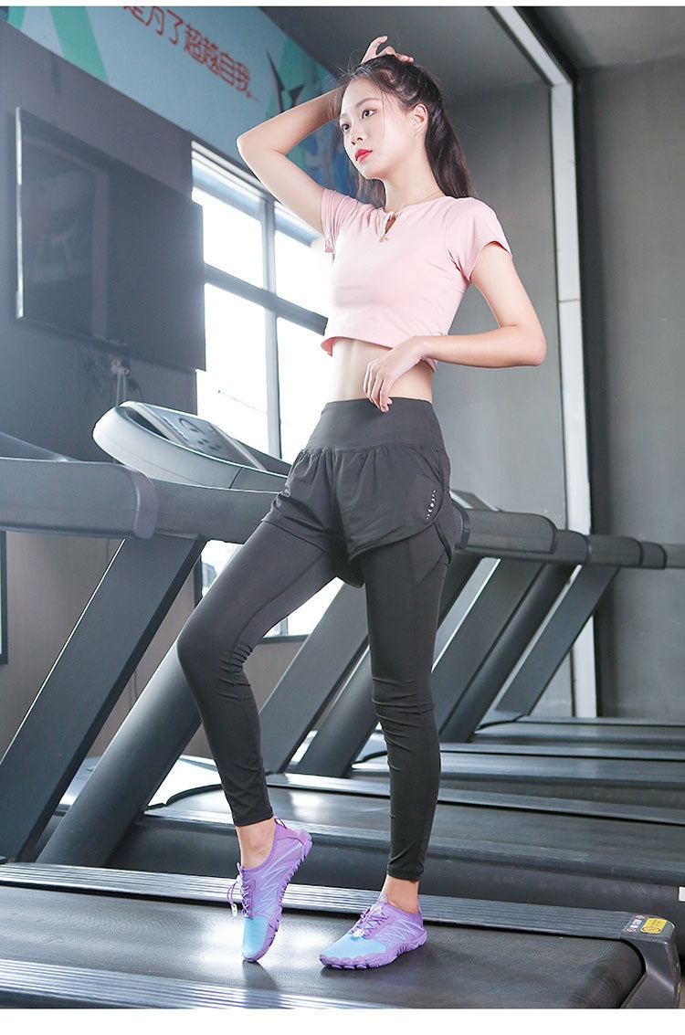 A woman posing on a treadmill in a gym, wearing a light pink short-sleeve crop top, black high-waisted athletic leggings, and purple athletic sneakers.