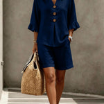 Woman wearing a navy blue outfit with shorts and a top, holding a straw bag against a neutral background.
