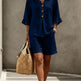 Woman wearing a navy blue outfit with shorts and a top, holding a straw bag against a neutral background.