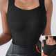 A woman wearing a black sleeveless scoop-neck bodysuit with built-in shoulder panels, paired with black faux-leather pants. The image includes a small inset showing the underbust area and an orange arrow indicating the shaping effect.