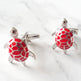 A pair of silver cufflinks shaped like small sea turtles, featuring bright red enamel scales on their shells, displayed on a light gray marble surface.