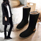 Black winter boots with fur lining on a wooden floor, with a person wearing a black coat in the background.