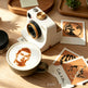 A white instant camera with gold accents sits on a wooden table beside a latte with latte art. Several printed photo cards are scattered nearby, including one that reads 'Cute kitti'.