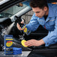 A man in a blue mechanic uniform cleans a car interior with a Ronavia plastic long-lasting restoration spray, using a yellow sponge and blue towels, with the product box shown in the foreground.
