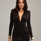 A woman modeling a black faux-leather long-sleeve bodycon mini dress with a plunging V-neckline and gold metallic cuffs and collar against a plain gray studio background.