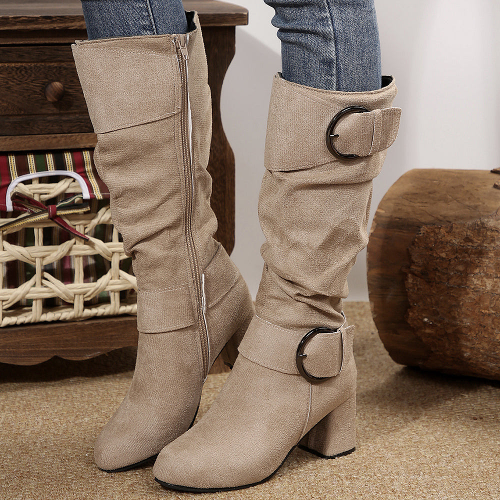 Beige knee-high boots with buckle details worn by a person, with a wooden stool and basket in the background.
