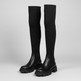 A pair of black over-the-knee boots with ribbed knit shafts, elastic side panels, and chunky lug soles, photographed on a light gray studio background.
