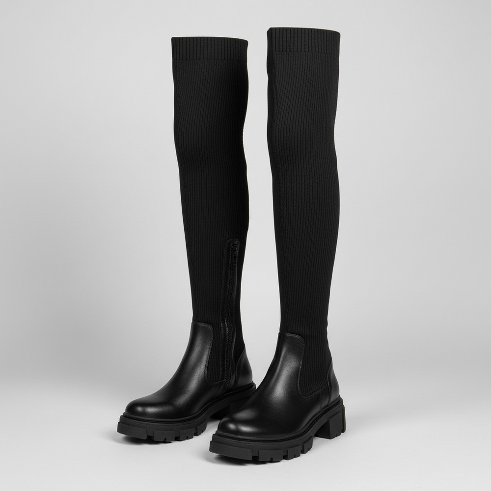 A pair of black over-the-knee boots with ribbed knit shafts, elastic side panels, and chunky lug soles, photographed on a light gray studio background.