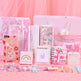 A pink-themed stationery gift set featuring notebooks, a glittery phone case, a correction tape dispenser, a small notebook, a sticker pad, and a pig-themed keychain, arranged on a pink surface with a pink tulle backdrop.