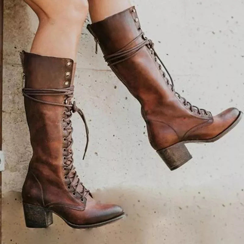 A pair of knee-high brown leather lace-up boots with block heels, worn on a person's feet against a light concrete floor.