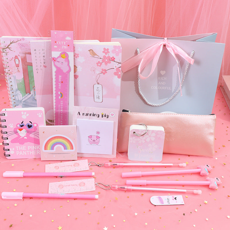 A pink-themed stationery gift set arranged on a pink backdrop with gold confetti. The set includes notebooks, pens, sticky notes, a small pouch, a card holder, and a large light blue gift bag with a pink ribbon.