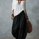 A woman modeling an off-the-shoulder white top and loose black harem-style pants, accessorized with layered necklaces, large hoop earrings, bracelets, and a woven handbag, against a textured gray studio backdrop.
