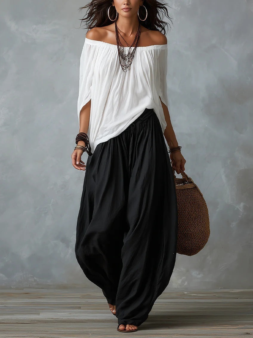 A woman modeling an off-the-shoulder white top and loose black harem-style pants, accessorized with layered necklaces, large hoop earrings, bracelets, and a woven handbag, against a textured gray studio backdrop.