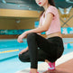 A woman crouches by an indoor swimming pool wearing a light pink short-sleeve crop top, black leggings, and bright pink and white running shoes.
