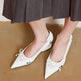 White pointed-toe shoes with bow details worn by a person on a brown floor.