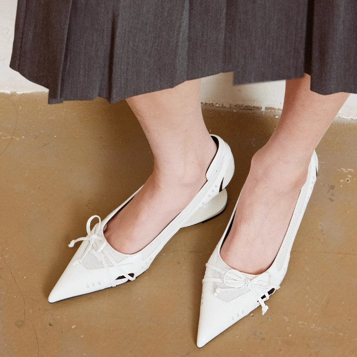 White pointed-toe shoes with bow details worn by a person on a brown floor.