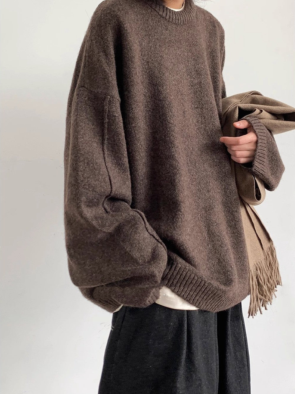 Person wearing a brown oversized sweater holding a beige coat against a white background
