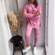 Person wearing a pink outfit standing in a room with a black stool and dried plants.