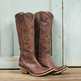 A pair of distressed brown leather cowboy boots with pointed toes, studded trim, and low stacked heels, displayed against a rustic light blue wooden background.