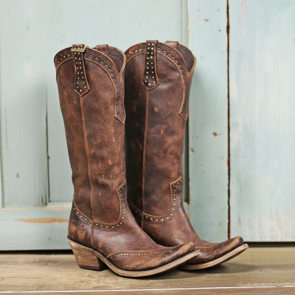 A pair of distressed brown leather cowboy boots with pointed toes, studded trim, and low stacked heels, displayed against a rustic light blue wooden background.