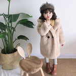 Child wearing a beige coat and hat standing next to a plant and wooden chair with rabbit ears.