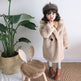 Child wearing a beige coat and hat standing next to a plant and wooden chair with rabbit ears.