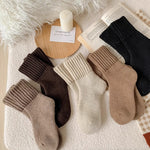 Four pairs of ribbed ankle socks arranged on a soft white textured surface, with decorative props including a candle, book, and fluffy white textile. The socks come in beige, brown, cream, and black.