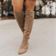 A person wearing knee-high taupe suede boots with a pointed toe and low block heel, standing on a light concrete surface outdoors.