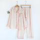 A two-piece pajama set consisting of a long-sleeve button-front top with a chest pocket and matching pants, both in a soft pink and off-white striped gauze fabric, hanging on wooden hangers against a plain white background.