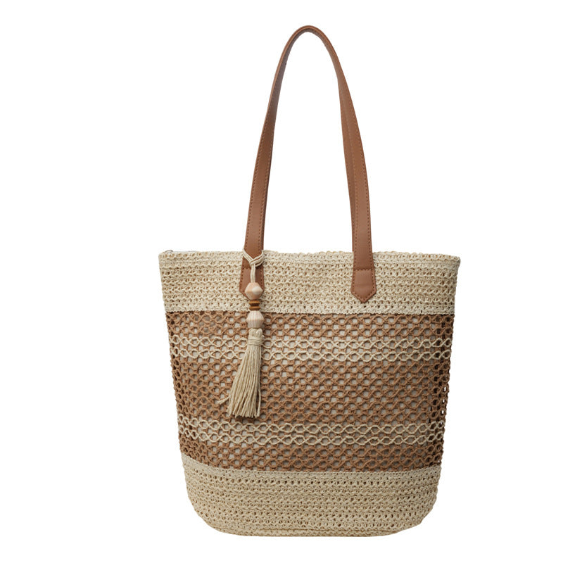 A woven straw tote bag with horizontal stripes in natural beige and medium brown, featuring tan leather shoulder straps and a matching tassel detail.