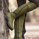 Olive green knee-high riding boots worn with dark brown leggings, shown on a person leaning against a tree outdoors.