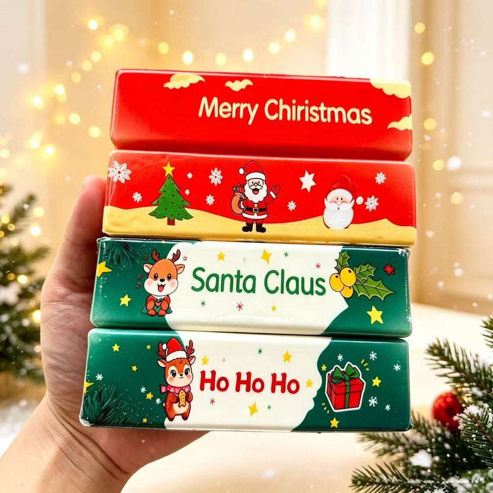 A hand holds a stack of four Christmas-themed rectangular boxes or packages decorated with festive illustrations and text including 'Merry Christmas,' 'Santa Claus,' and 'Ho Ho Ho.' The background is softly blurred with warm lights and holiday greenery.