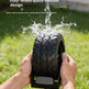 A person holds a black tire-shaped cleaning brush or roller outdoors over running water, with grass and a blurred outdoor background. White text at the top reads 'Four-wheel quick-release design' and 'Cleans without dead corners, easily removes dirt from crevices.
