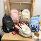 Five backpacks in various colors on a wooden shelf. Large Capacity Nylon Backpack – Cartoon Cute Style | 10 Colours