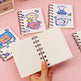 A hand holds an open spiral-bound notebook with blank cream pages, surrounded by other closed notebooks featuring cute cartoon space-themed illustrations on a pink background.