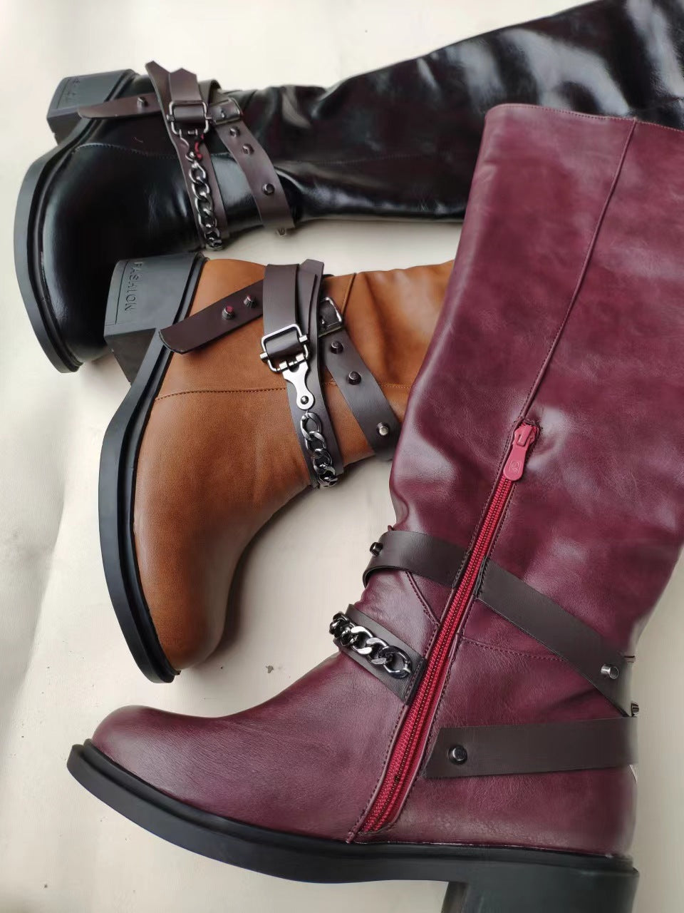 Three boots in black, brown, and burgundy with decorative straps and zippers on a light background.