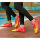 A close-up of a person's legs wearing bright red and yellow athletic water shoes with black leggings, standing on a wet wooden deck next to a swimming pool.