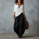 Woman wearing a white off-shoulder top and black harem pants, holding a woven bag against a gray background.