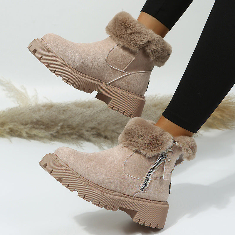 A pair of women's ankle boots in light taupe suede with fluffy faux-fur trim and a side zipper, shown being worn with black leggings. The boots feature chunky lug soles and are photographed against a light studio background with a soft beige pampas grass prop.