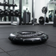 A black grip strengthener hand grip on a dark gray rubber gym floor, with blurred weight racks and dumbbells in the background.