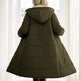 A woman viewed from behind wearing a long olive green hooded puffer coat in a bright room with white curtains and a window.