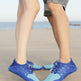 A close-up of two people standing on a sandy beach wearing blue and light blue minimalist water shoes.
