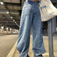 Person wearing blue jeans and a light blue shirt, standing on a sidewalk with a white bag.