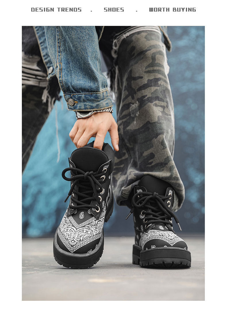 A person wearing black and white bandana-patterned lace-up boots, with one boot being held by a hand. The person is also wearing gray camouflage pants and a blue denim jacket, set against a textured blue background.